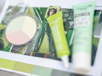Nahaufnahme von Make-up-Produkten, darunter eine runde Puderdose mit Pastelltönen und zwei grünen Tuben, vor einem bunten Hintergrund.