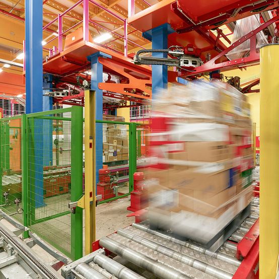 Ein farbenfrohes Logistikzentrum mit einem Fördersystem, das einen unscharfen Stapel von Kisten bewegt, umgeben von grünen Sicherheitsgeländern und lebendigen Maschinen.