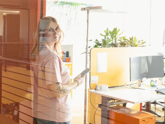 Frau mit Tattoos, die ein Telefon in der Hand hält, steht in einem hellen Büro in der Nähe eines Schreibtisches mit einem Computer und Pflanzen und schaut durch eine Glaswand.