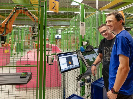 Zwei Personen stehen in einem Logistikzentrum neben Computermonitoren, die Daten anzeigen, während im Hintergrund Maschinen und Sicherheitszäune zu sehen sind.