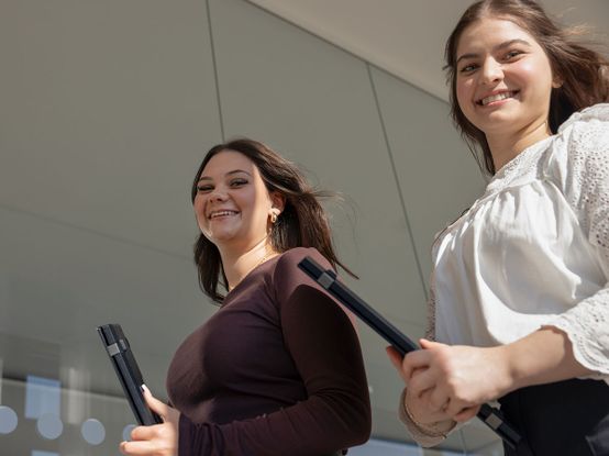 Zwei Frauen, die lächelnd und mit Laptops in der Hand durch ein helles, modernes Gebäude mit Glaswänden gehen.