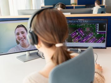 Person mit Kopfhörern, die an einem Schreibtisch mit zwei Monitoren sitzt; auf dem einen wird ein Videoanruf angezeigt, auf dem anderen eine technische Zeichnung.