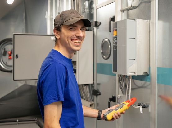 Eine lächelnde Person in blauem Hemd und Mütze hält ein Werkzeug in der Hand und steht neben einer Industrieanlage in einem technischen Umfeld.