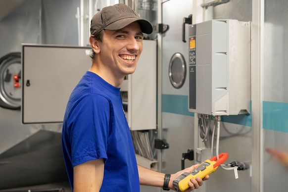 Eine lächelnde Person in blauem Hemd und Mütze hält ein Werkzeug in der Hand und steht neben einer Industrieanlage in einem technischen Umfeld.