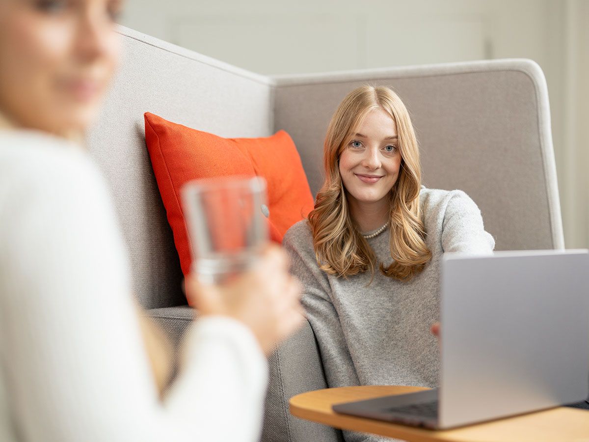 Zwei Menschen in einer gemütlichen Umgebung, einer mit einem Glas in der Hand, der andere lächelnd am Laptop, mit einem orangefarbenen Kissen im Hintergrund.
