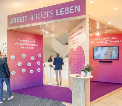 Ausstellungsstand in rosa und Holzdesign mit interaktiven Displays und Text über das Arbeiten bei dm-drogerie markt. Die Menschen beschäftigen sich mit den Displays.