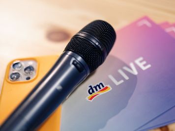 Ein schwarzes Mikrofon liegt auf einem Stapel Moderationskarten mit der Aufschrift „LIVE“, daneben ein orangefarbenes Smartphone auf einer Holzfläche.