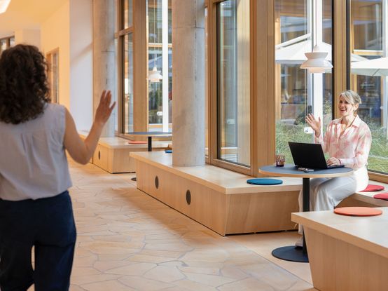 Eine Frau, die an einem Tisch mit einem Laptop sitzt, winkt einer anderen Frau zu, die in einem Flur mit großen Fenstern vorbeigeht.