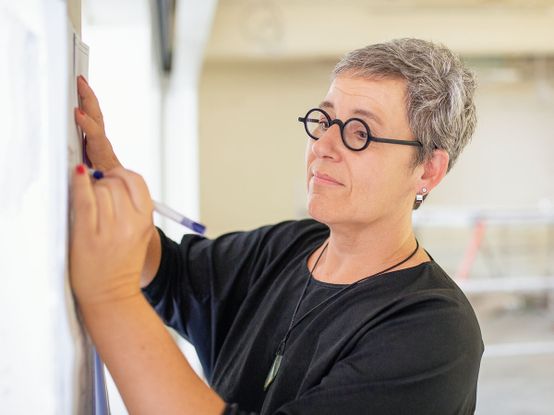Person mit kurzen grauen Haaren und runder Brille, die in einem hellen Innenraum mit einem Stift auf einen Bauplan schreibt.