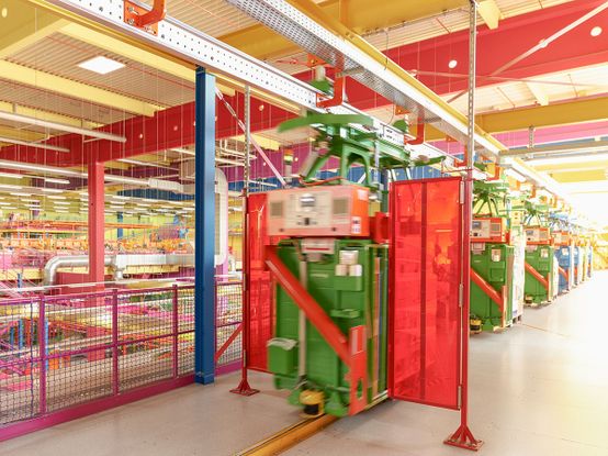 Buntes Logistikinterieur mit Hängebahnen, darunter grüne und rote Geräte auf Schienen, umgeben von gelben und rosa Strukturelementen.