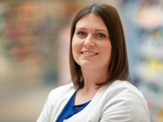 Eine Frau mit schulterlangem braunem Haar lächelt in einem blauen Oberteil und einer weißen Jacke vor einem unscharfen Hintergrund.