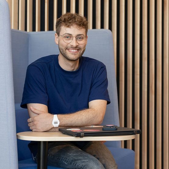 Lächelnder Mann mit Brille und blauem Hemd sitzt in einer modernen Lounge mit einem kleinen Tisch und einem Hintergrund aus Holzlatten.