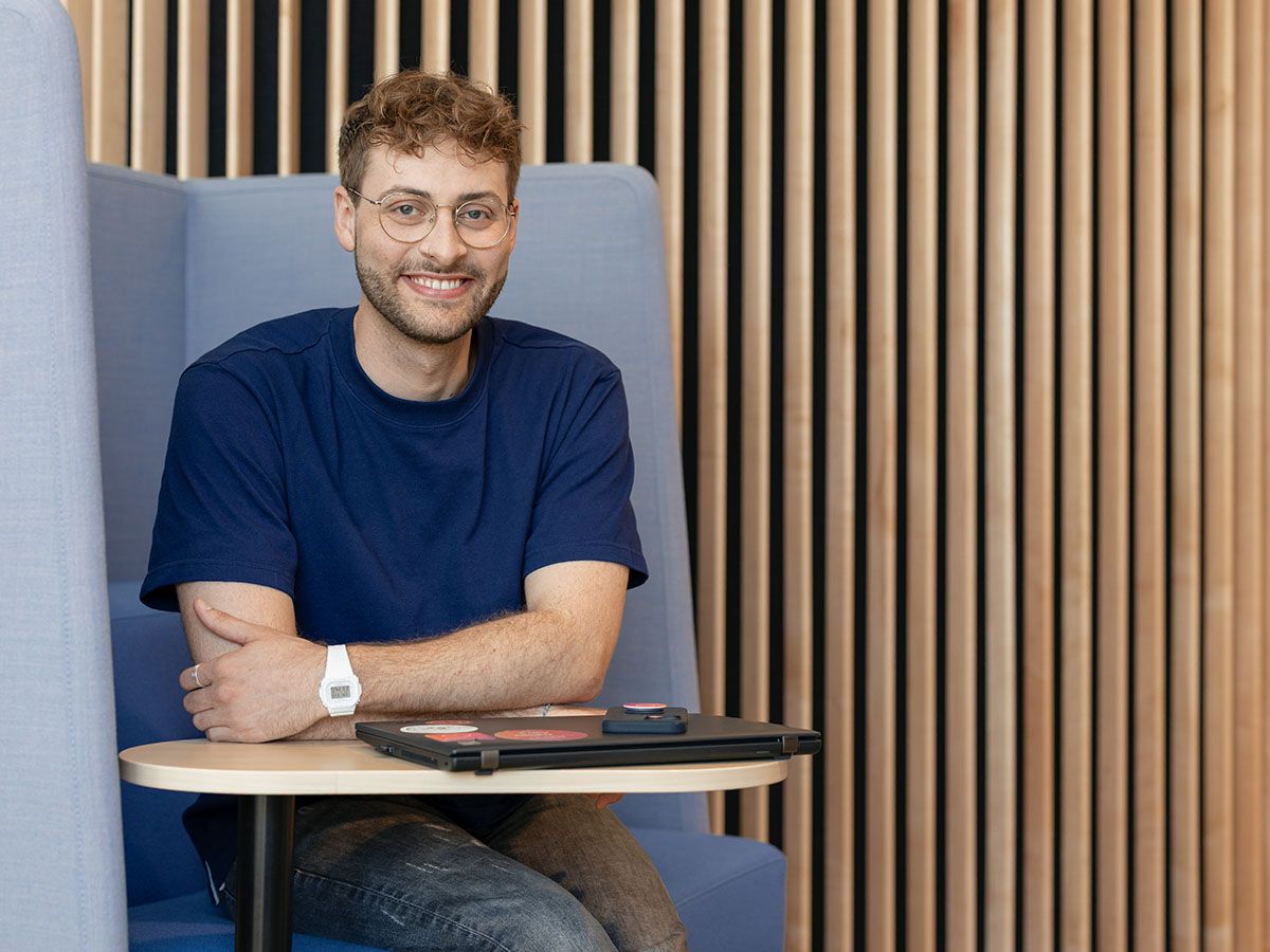 Lächelnder Mann mit Brille und blauem Hemd sitzt in einer modernen Lounge mit einem kleinen Tisch und einem Hintergrund aus Holzlatten.