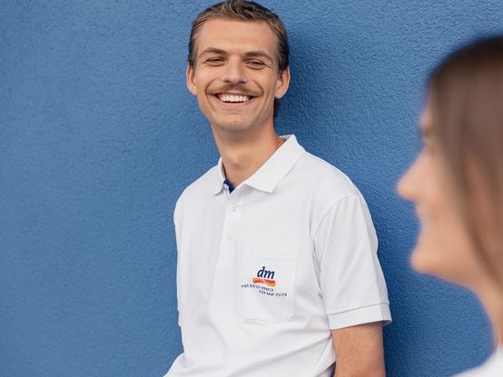 Ein lächelnder Mann in einem weißen Polohemd mit dm-Logo, der an einer blauen Wand lehnt, blickt in Richtung einer verschwommenen Person im Vordergrund.