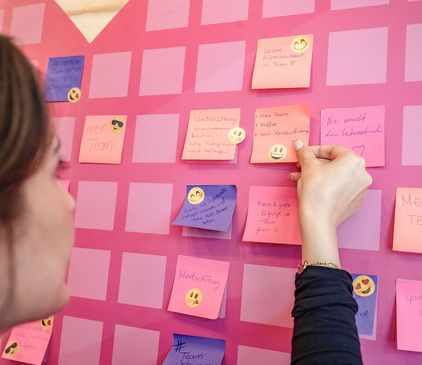 Frau ordnet bunte Haftnotizen mit Emojis auf einem rosa Gitterbrett an.