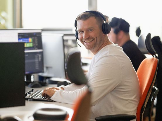 Mann im weißen Hemd und mit Kopfhörern, lächelnd an einem Schreibtisch mit mehreren Computermonitoren in einer Büroumgebung.
