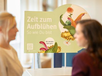Zwei Personen stehen neben einem Schild mit Blumengrafik und deutschem Text: "Zeit zum Aufblühen. So wie Du bist. Wir beraten Dich gerne."