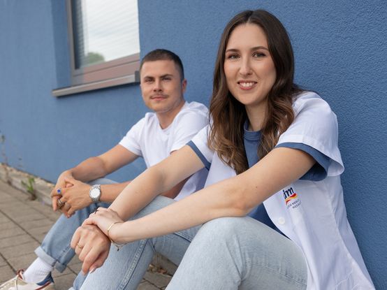 Zwei Personen sitzen vor einer blauen Wand; die Frau trägt einen weißen Kasack und lächelt, während der Mann ein weißes T-Shirt trägt und entspannt im Hintergrund sitzt.