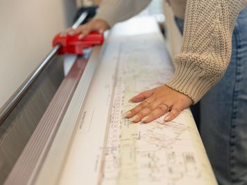 Person, die mit einem großen Papierschneider einen Architekturplan zurechtschneidet, in einem beigen Pullover und Jeans.
