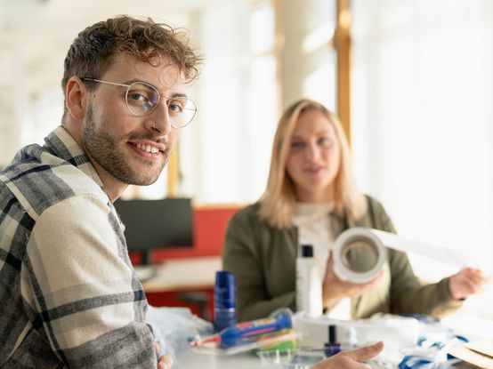 Ein Mann mit Brille lächelt in die Kamera, während eine Frau in einer hellen Büroumgebung eine Rolle mit Fotopapier hält. Verschiedene Büromaterialien liegen auf dem Tisch.