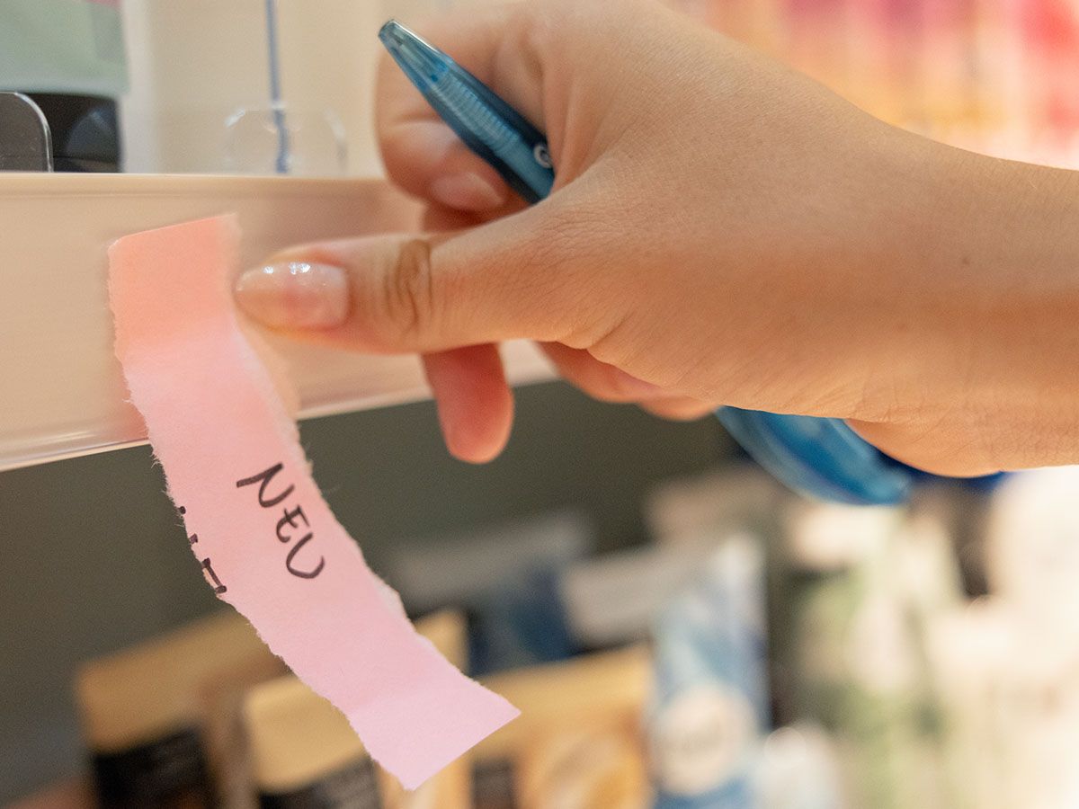 Hand, die ein rosafarbenes Papier mit der Aufschrift „NEU“ auf ein Regal legt, mit unscharfen Produkten im Hintergrund.