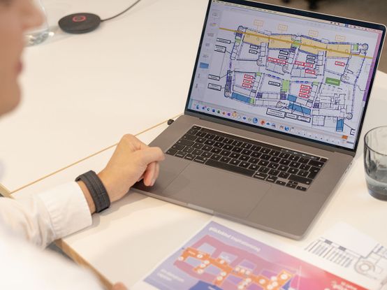Person mit einem Laptop, der einen bunten Grundriss zeigt, mit einem gedruckten Layout und einem Glas Wasser auf dem Tisch.