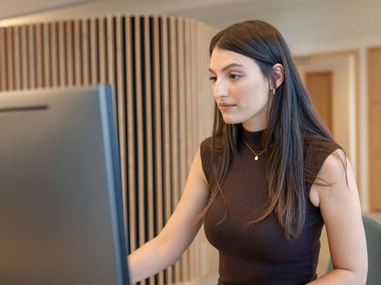 Frau mit langen braunen Haaren arbeitet an einem Computer in einem modernen Büro mit Holzverkleidung im Hintergrund.