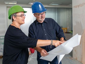 Zwei Personen mit Schutzhelmen prüfen auf einer Baustelle Architekturpläne. Einer zeigt auf ein Detail auf dem Papier.