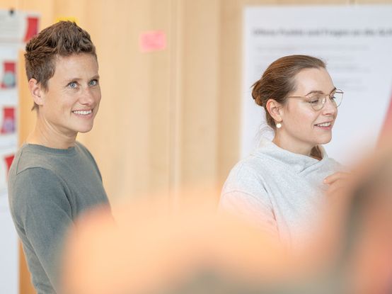 Zwei lächelnde und sich unterhaltende Personen in einem hellen Raum, mit unscharfen Vordergrundelementen. Eine trägt eine Brille, die andere hat kurzes Haar.