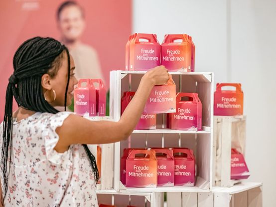 Eine Frau wählt eine bunte Schachtel mit der Aufschrift „Freude zum Mitnehmen“ aus einer Reihe ähnlicher Schachteln auf weißen Regalen aus.