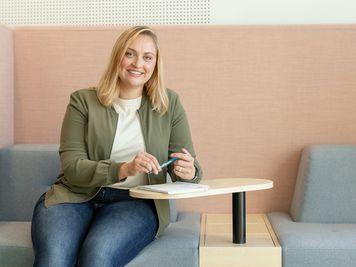 Lächelnde Frau, die auf einer modernen Couch sitzt, einen Stift und ein Notizbuch in der Hand hält, eine grüne Jacke und Jeans trägt und einen hellrosa Hintergrund hat.