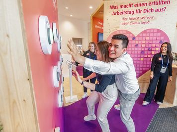 Zwei Personen spielen mit einer Wand aus Knöpfen an einem Veranstaltungsstand. Andere schauen zu und lächeln. Der Stand zeigt einen Text auf Deutsch.