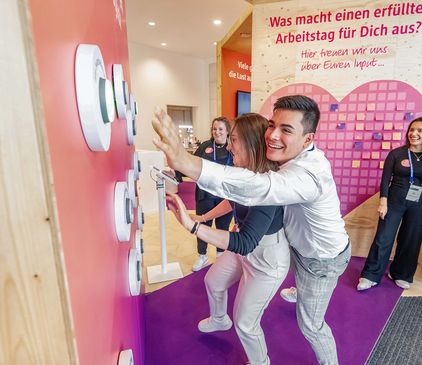 Zwei Personen spielen mit einer Wand aus Knöpfen an einem Veranstaltungsstand. Andere schauen zu und lächeln. Der Stand zeigt einen Text auf Deutsch.