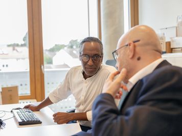 Zwei Männer in einem Büro, einer im weißen Hemd am Computer, der andere im Anzug, unterhalten sich am Schreibtisch.