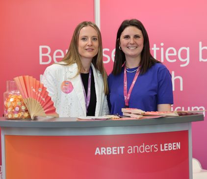 Zwei junge Frauen lächeln hinter einem Counter mit dem Schriftzug „Arbeit anders leben“